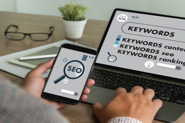 Person uses a smartphone with an SEO magnifying glass icon while a laptop screen shows keywords and SEO ranking concepts.