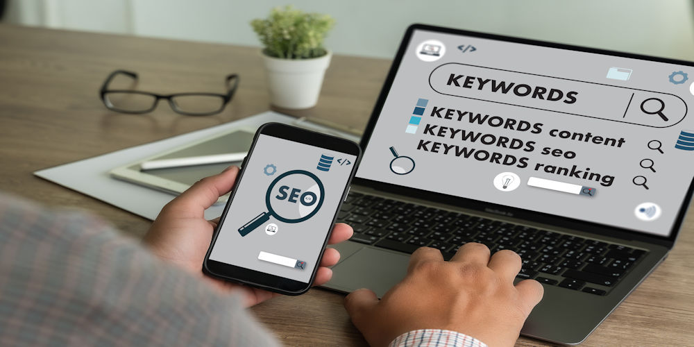 Person uses a smartphone with an SEO magnifying glass icon while a laptop screen shows keywords and SEO ranking concepts.