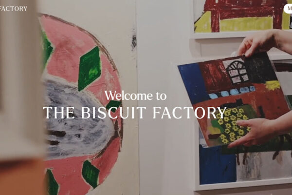 Person holding colorful abstract paintings in an art gallery or studio; overlay text reads 'Welcome to THE BISCUIT FACTORY'