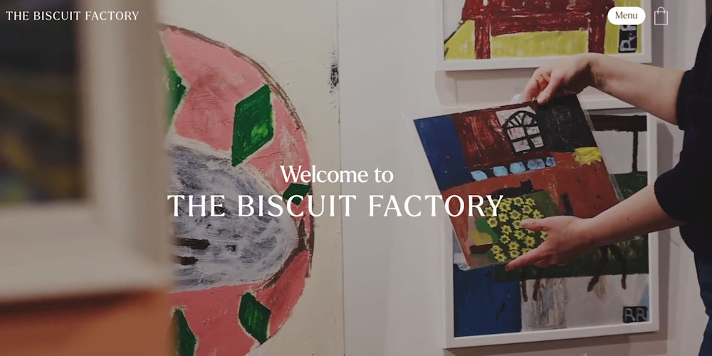 Person holding colorful abstract paintings in an art gallery or studio; overlay text reads 'Welcome to THE BISCUIT FACTORY'