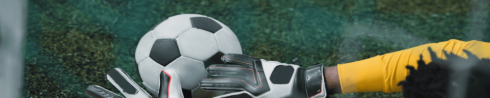 sportsshirts-1000-1 - Marketing North East Soccer goalkeeper's gloved hands reaching to stop a black-and-white soccer ball on green turf field.