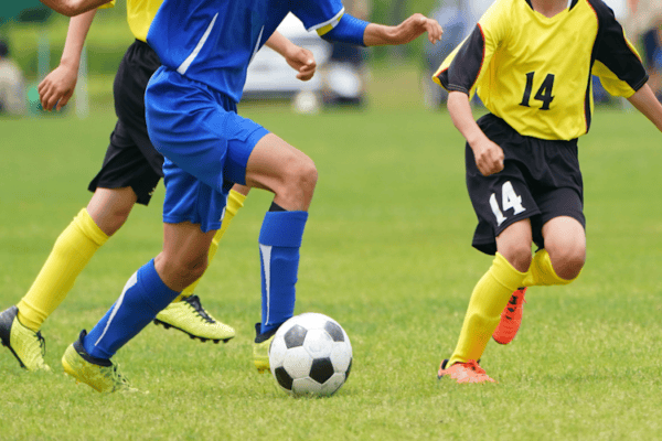 Young soccer players in blue and yellow kits chase a soccer ball on a grassy field during a youth match.