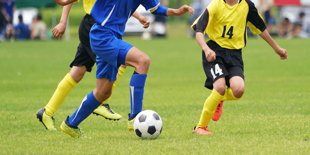 Young soccer players in blue and yellow kits chase a soccer ball on a grassy field during a youth match.